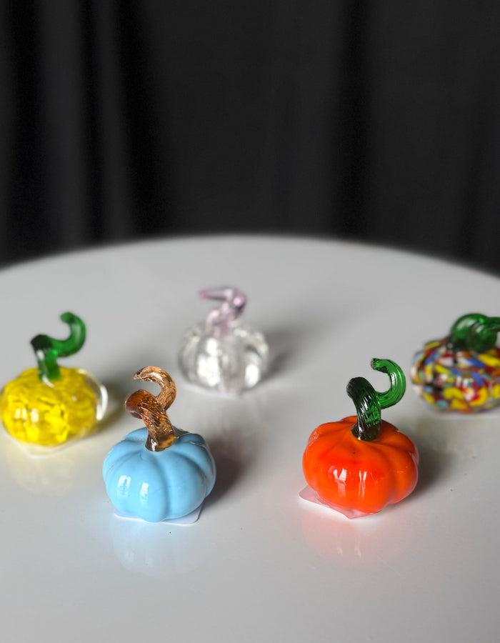 Tiny glass pumpkins with blue and orange pumpkins in the foreground and yellow, multicolored, and clear pumpkins behind.